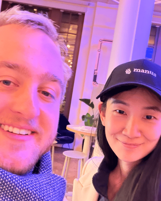 A close-up selfie taken indoors under dramatic pink and purple lighting, featuring a man and a woman smiling at the camera. The woman is wearing a dark baseball cap with the word 'manus' embroidered on it.