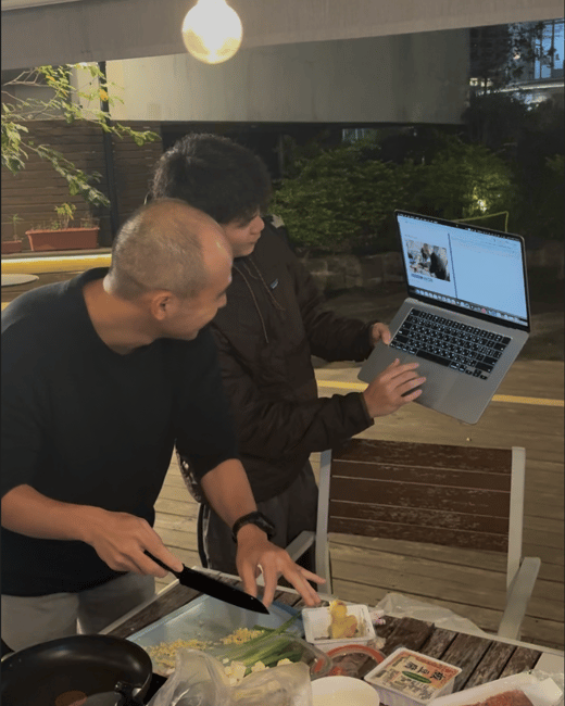 Two men are standing around an outdoor table at night, preparing food; one man chops ingredients while the other holds up a laptop, possibly viewing a recipe or video call.