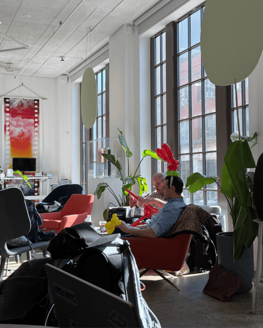 A bright, modern indoor lounge or office space where two men are sitting and interacting with balloon animals near large windows.