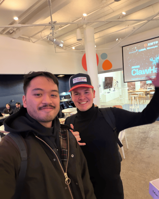 Two men posing for a selfie in an indoor event space, with a projector screen displaying hackathon details in the background.