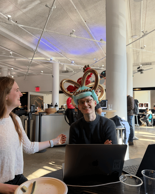 A man wearing an elaborate balloon crown sits at a table with a laptop, smiling at the camera, while a woman next to him laughs in a modern indoor setting.
