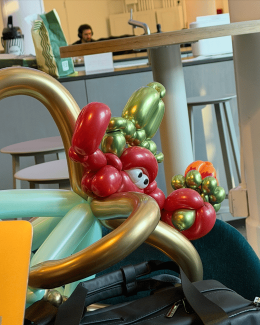 A vibrant balloon sculpture of a character with large eyes sits on a teal chair next to a black bag in a modern indoor setting. A person is visible in the background sitting at a table.