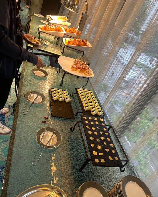 A buffet table is elegantly set with various appetizers, including fried chicken drumsticks, skewers, and small pastries, while a person reaches for a snack.