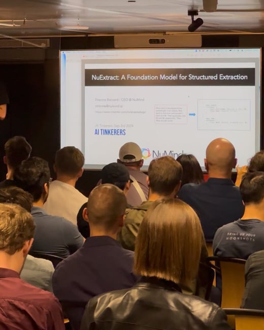 A photograph showing a large audience seated in a dimly lit room, viewing a presentation slide on a projector screen during an event focused on AI and technology.