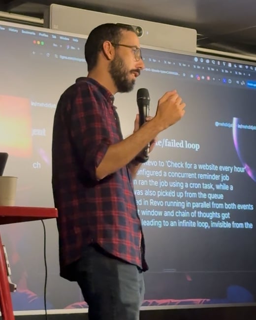 A man wearing a plaid shirt, glasses, and a beard is giving a technical presentation indoors while holding a microphone and gesturing with his hand. Behind him, a large screen displays white text on a dark background detailing a 'failed loop' scenario.