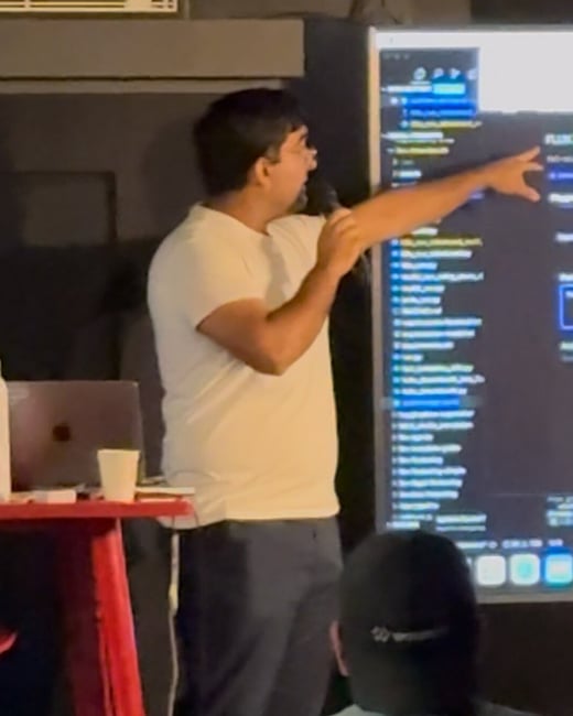 A man is giving a technical presentation in a dimly lit room, holding a microphone and pointing at a large vertical screen displaying lines of code or technical file structures.