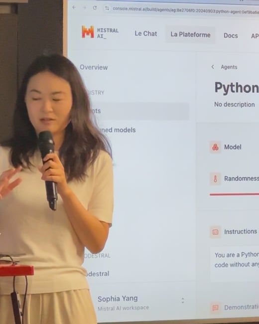 A woman, identified as Sophia Yang, is giving a presentation while holding a microphone, standing in front of a large screen displaying the Mistral AI console interface.