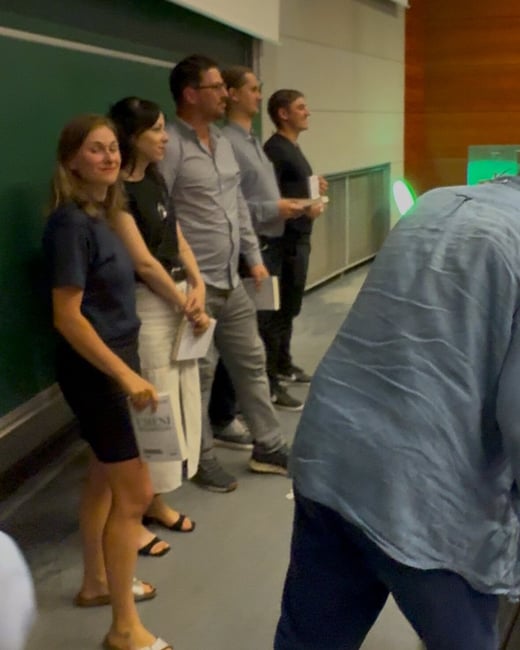 A candid, low-quality photograph capturing a line of five people (two women and three men) standing against a dark green wall, likely in a lecture hall or conference setting. A sixth person in a blue shirt is heavily blurred and obscures the lower right foreground.