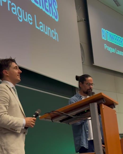 A photo taken during a professional event, showing two men on a stage in a lecture hall setting, with presentation slides visible in the background displaying the text 'AITINKERERS Prague Launch.'