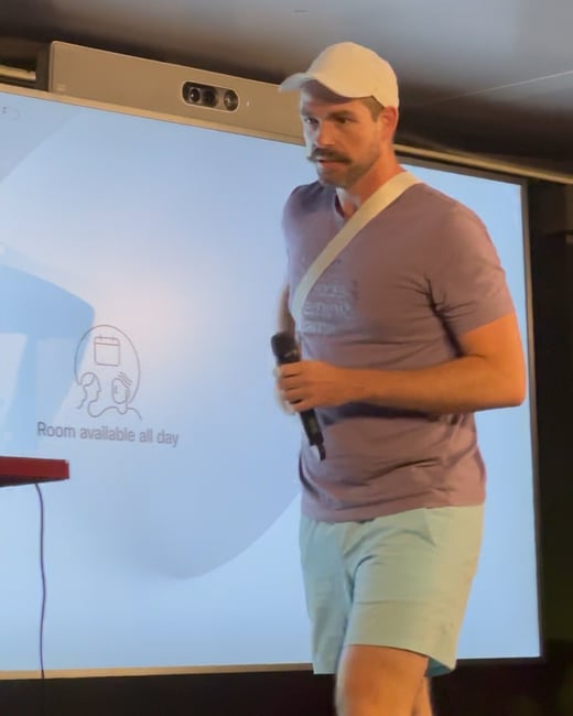 A man with a prominent mustache, wearing a white cap, purple t-shirt, and light blue shorts, stands in front of a large screen holding a microphone, likely giving a casual presentation or speech.