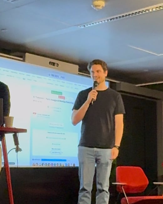 A man in a black t-shirt and jeans is standing on a stage, holding a microphone and giving a presentation in front of a large projection screen.