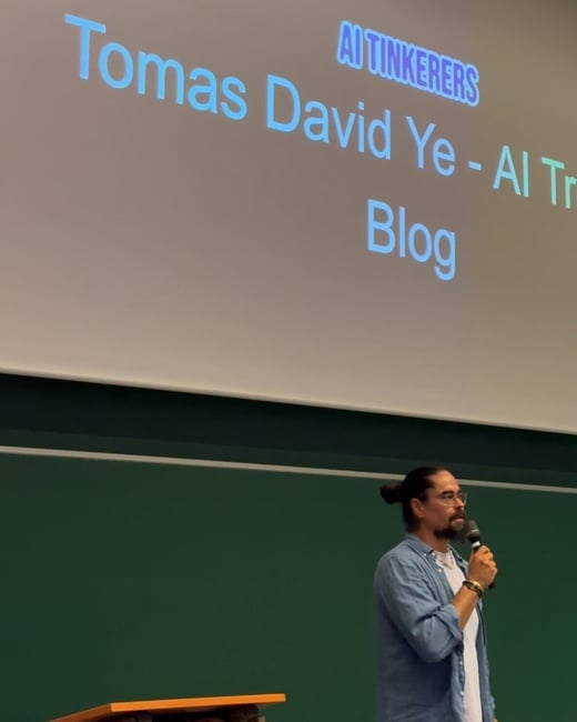 A photograph showing a man speaking into a microphone during a presentation, standing in front of a large screen displaying the text, 'Tomas David Ye - Al Tr[unreadable] Blog' and 'AI TINKERERS'.