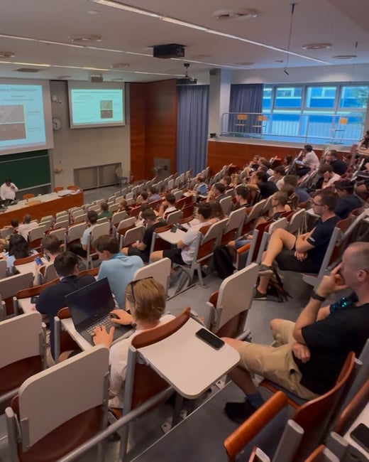A photograph capturing a lecture or presentation in a large, tiered university auditorium filled with students. A speaker stands at the front near a podium, and a technical slide is projected onto a screen.