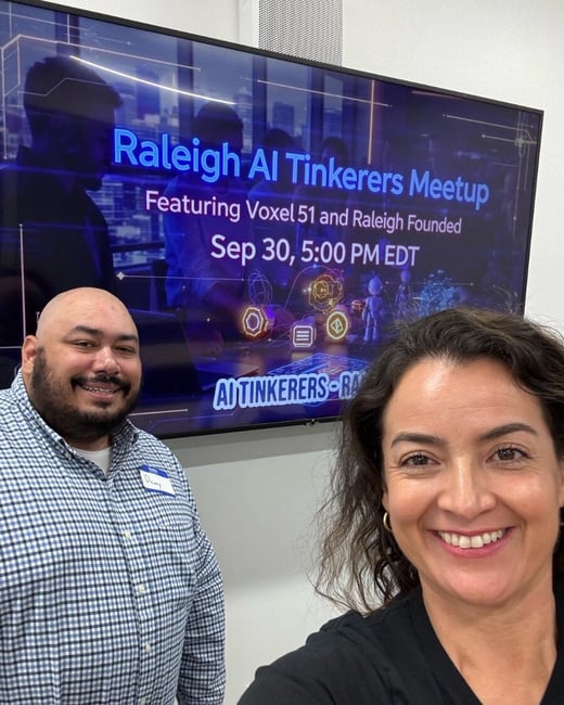 A smiling selfie of a woman and a man taken indoors, with a large screen visible behind them advertising the "Raleigh AI Tinkerers Meetup."