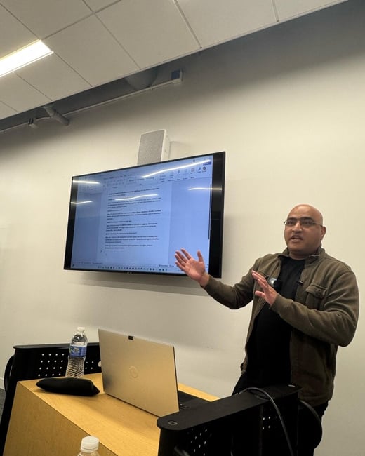 A man wearing glasses and a brown jacket is giving a presentation or lecture in a modern classroom, standing next to a large wall-mounted monitor displaying a text document.