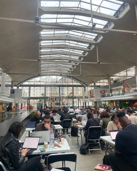 A wide-angle photograph captures a large, modern indoor hall with a high, vaulted concrete and glass ceiling, where numerous people are seated at white folding tables working intently on laptops.