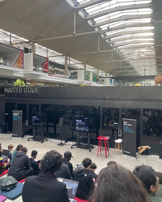 A photograph capturing a large indoor tech event, likely a hackathon, where an audience is seated on the floor facing the 'MASTER STAGE' which features two screens displaying 'Paris AI Hackathon.'
