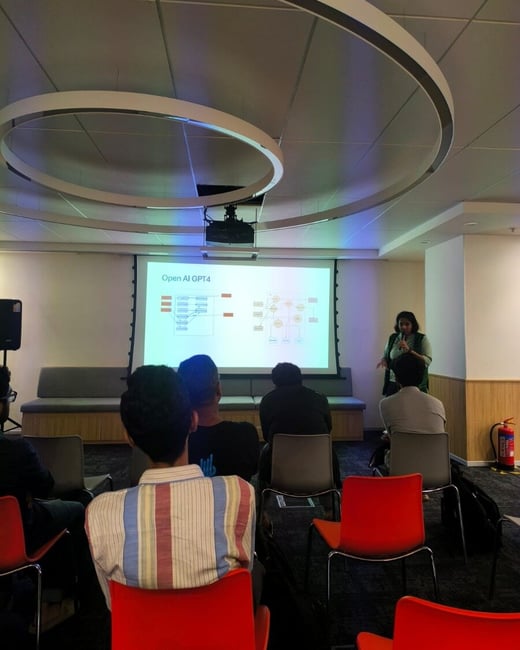A photograph captures an indoor presentation where a woman is speaking to an audience next to a projector screen displaying a diagram titled "Open AI GPT4."