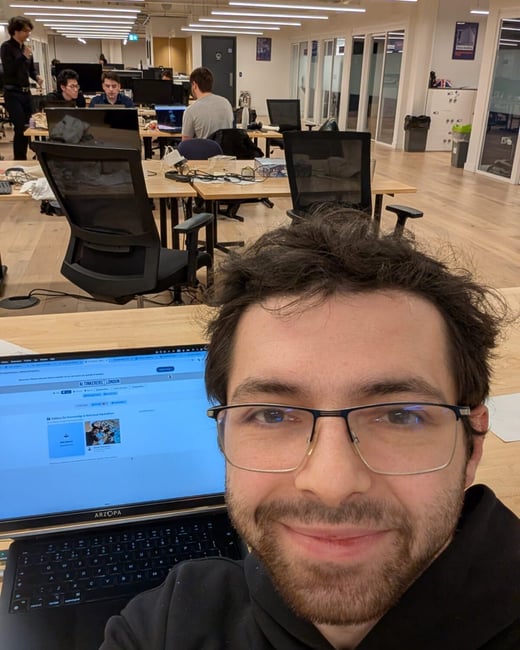 A man wearing glasses and a black hoodie takes a close-up selfie while sitting at his desk in a brightly lit, open-plan office where several other people are working in the background.