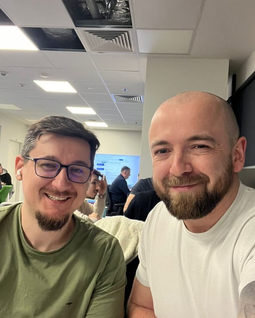 A close-up photograph of two smiling men, one wearing glasses and an olive green shirt and the other a white shirt, taken in a brightly lit indoor office environment with other colleagues visible in the background.
