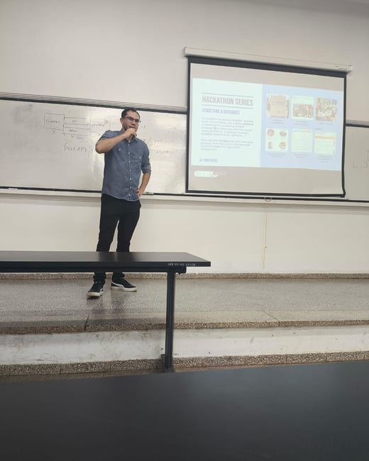 A man stands at the front of a room, giving a presentation with a whiteboard and a projection screen behind him.