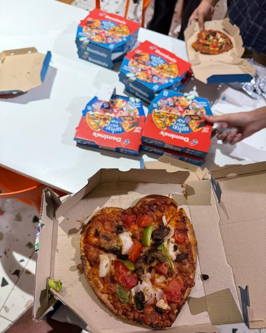 A heart-shaped pizza sits in an open box in the foreground, with several more Domino's pizza boxes and people's hands visible on a white table in the background.