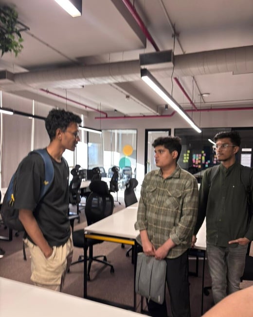 Three young men are standing and talking in a modern, open-plan office space with white desks and industrial-style ceilings.
