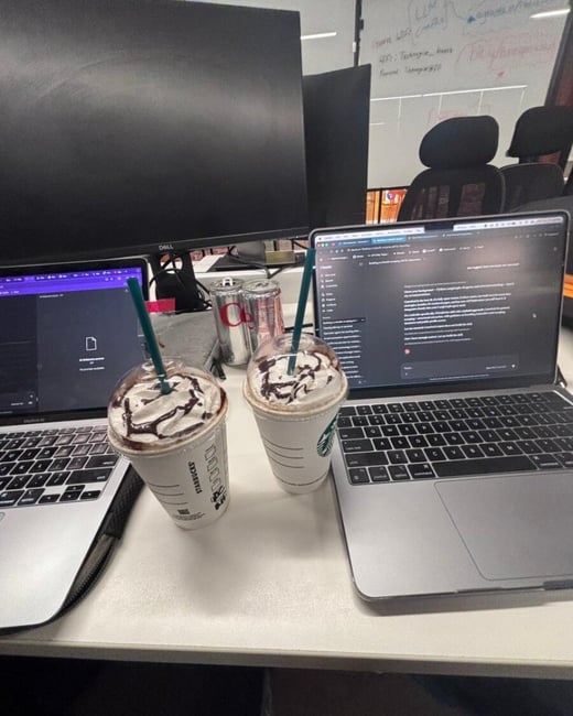 A workspace setup featuring two Starbucks frappuccinos placed between two open laptops on a white desk, with a large monitor and a whiteboard in the background.