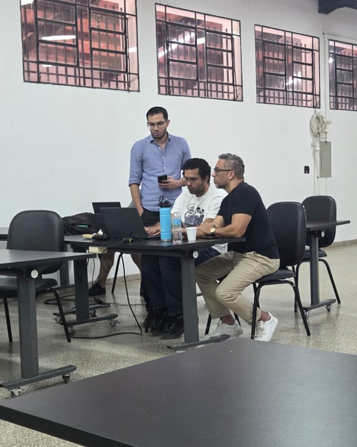 Three men are gathered around a desk with laptops in an indoor space, likely a classroom or office setting.