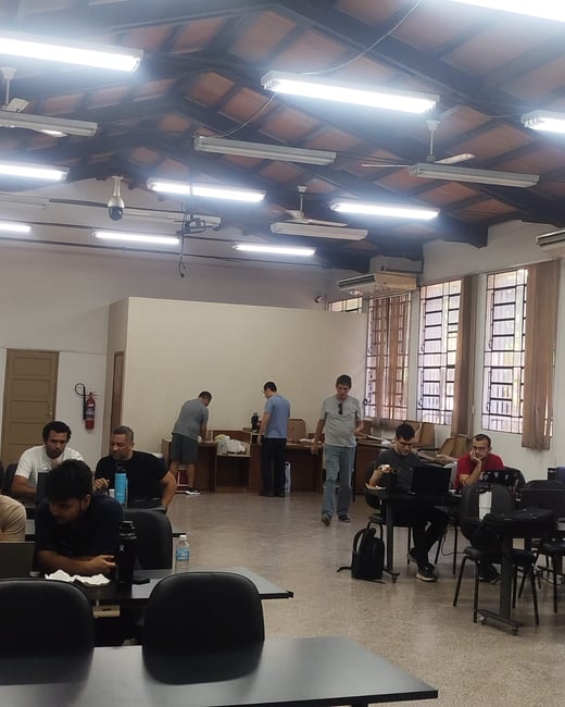 A group of people are gathered in a large indoor workspace with high ceilings and exposed beams, some working on laptops while others stand in the background.