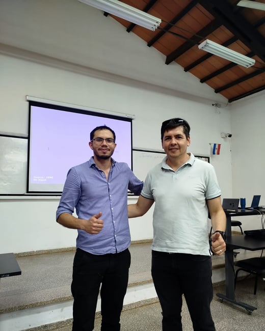 Two men are standing in a classroom or presentation room, smiling and giving a thumbs-up gesture in front of a projector screen.
