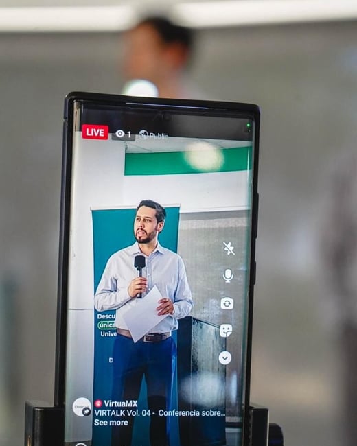 A smartphone mounted on a tripod is live-streaming a man giving a presentation with a microphone and notes in hand.