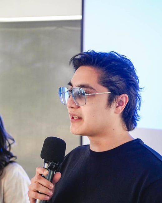 A young person with dark hair and glasses is captured in a candid moment, speaking into a microphone while holding a piece of paper.