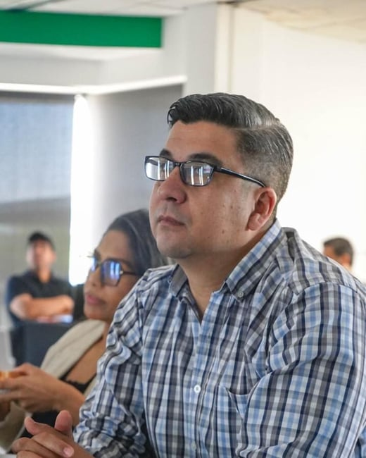 A man wearing glasses and a plaid shirt is sitting at a table with a notebook, looking attentively towards something off-camera during a meeting.