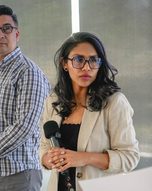 A woman with dark hair and glasses holds a microphone during an indoor event, while a man in a plaid shirt stands beside her.