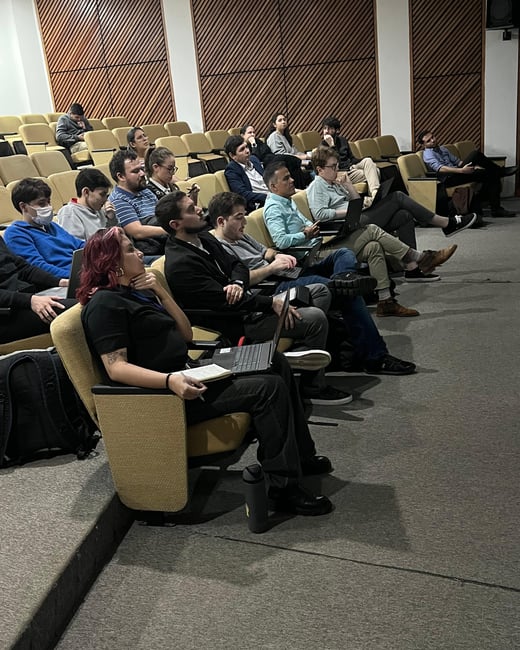 An audience of people sitting in an auditorium, many of whom are using laptops and taking notes while looking toward the front of the room.