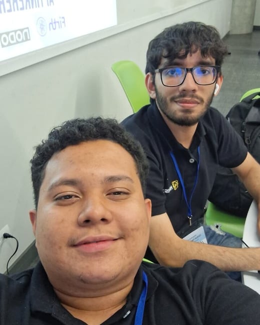 A selfie of two young men wearing black polo shirts and blue lanyards in an indoor setting, possibly a classroom or office.