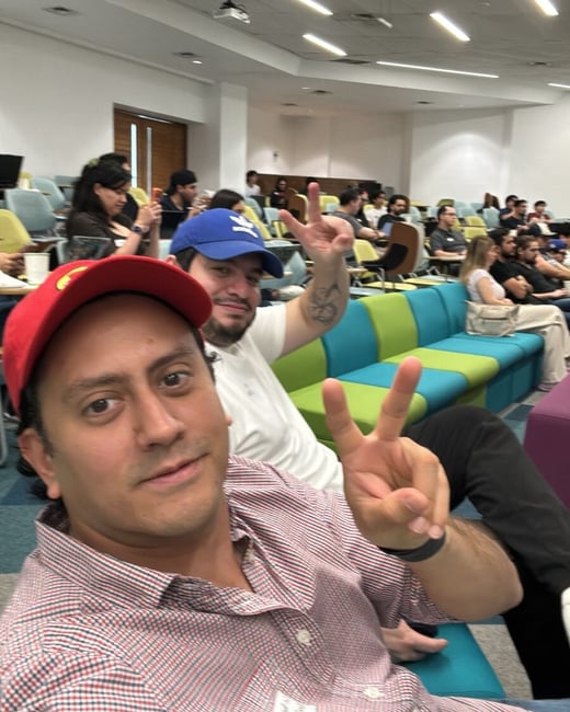A selfie of two men making peace signs in a large lecture hall or auditorium filled with an audience.
