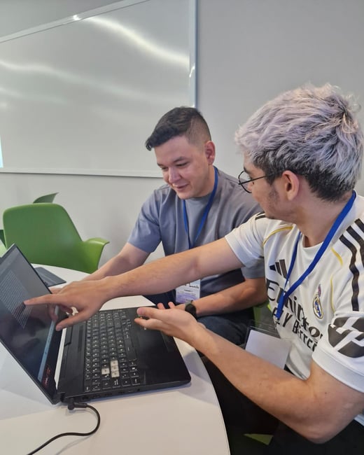 Two men are sitting at a table indoors, focused on a laptop screen while one points at the display.