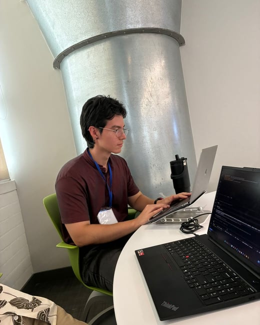 A man with glasses and a maroon shirt is sitting at a desk, working on a laptop. An industrial metal vent is visible in the background.