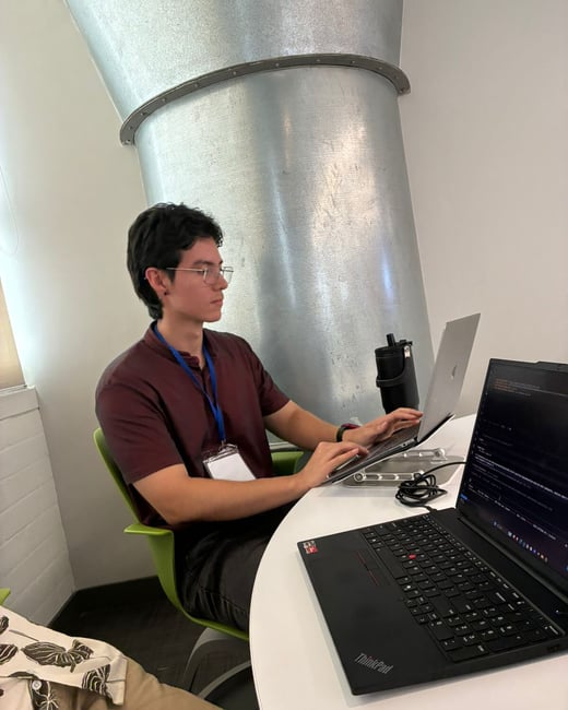 A young man wearing glasses and a maroon shirt is sitting at a white table, working on two laptops in an indoor setting with a large metal duct in the background.
