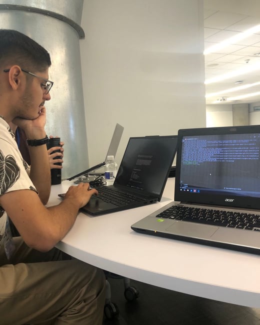 A man in glasses sits at a white table, focused on a laptop screen showing terminal error messages in an office setting.