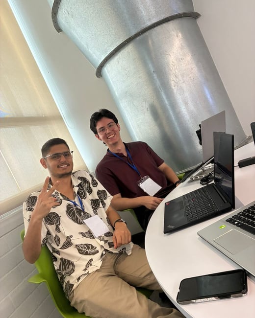 Two young men are sitting at a white table with laptops, smiling and posing for a photo in an indoor setting.