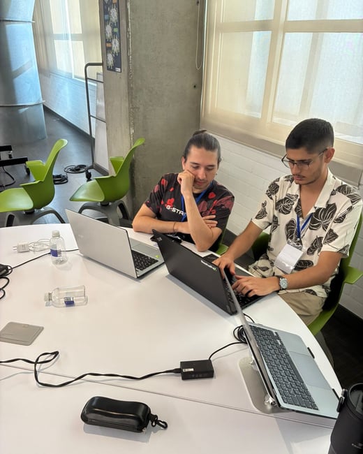 Two men are sitting at a white table in a brightly lit indoor space, focused on their laptops.