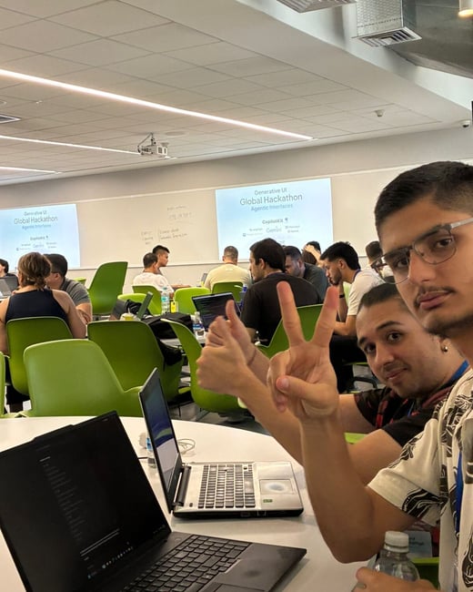 A group of people participating in a hackathon event, with two men in the foreground making hand gestures toward the camera while sitting at a table with laptops.