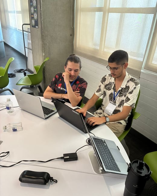 Two men are sitting at a white table in an office setting, focused on their laptops and collaborating.