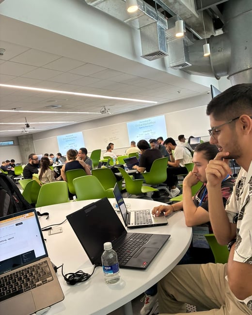 A group of people are working on laptops at round tables in a modern, open-plan room during a hackathon event.