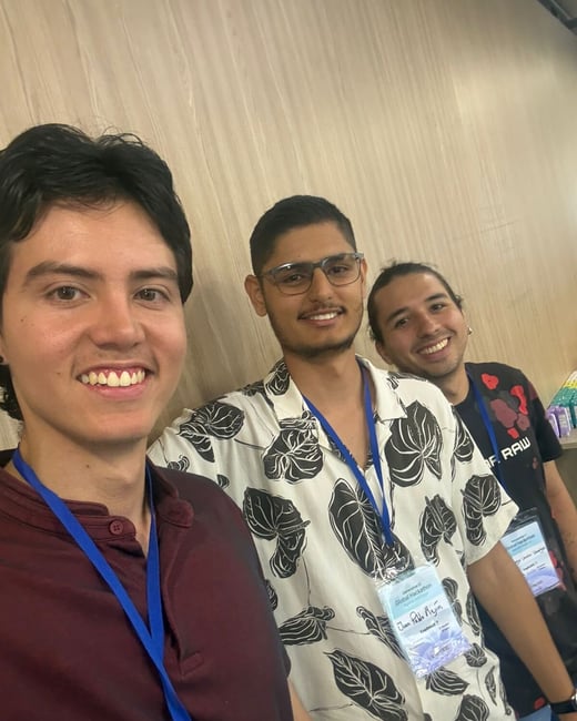 Three young men are smiling for a selfie at what appears to be a hackathon event, wearing lanyards with identification badges.