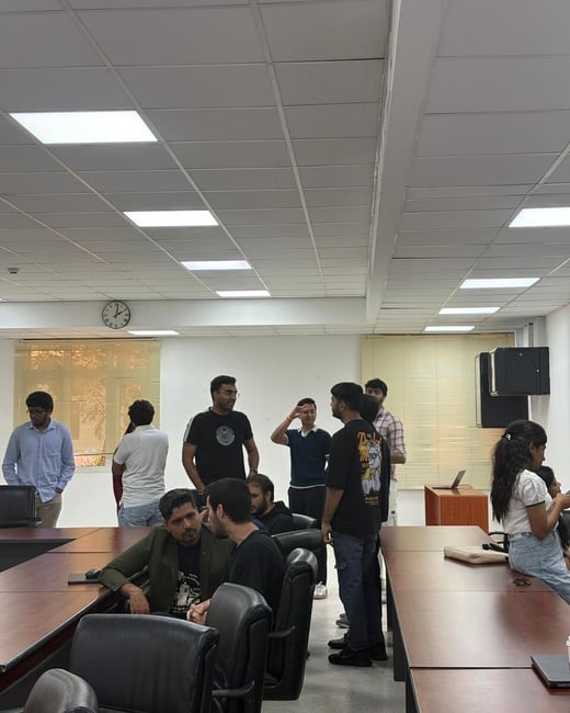 A group of people are gathered in an office or classroom setting, standing and sitting around conference tables while conversing.