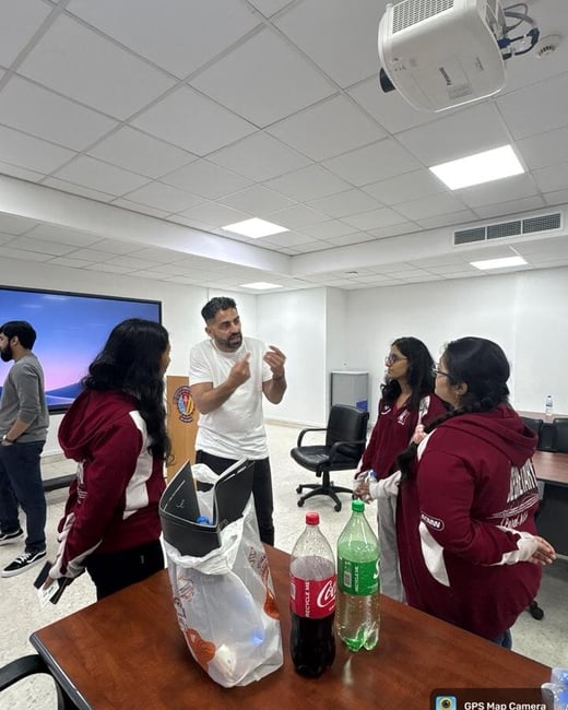 A group of individuals in maroon hoodies are engaged in a discussion with a man in a white t-shirt inside a classroom or meeting room.